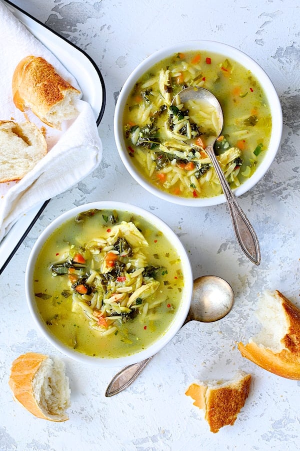 Photo of two bowls of Cold-Fighting Chicken Soup with Kale and Orzo with bread scattered around.