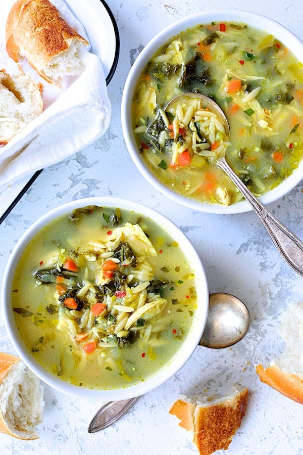 Photo of two bowls of Cold-Fighting Chicken Soup with Kale and Orzo with bread scattered around the bowls.