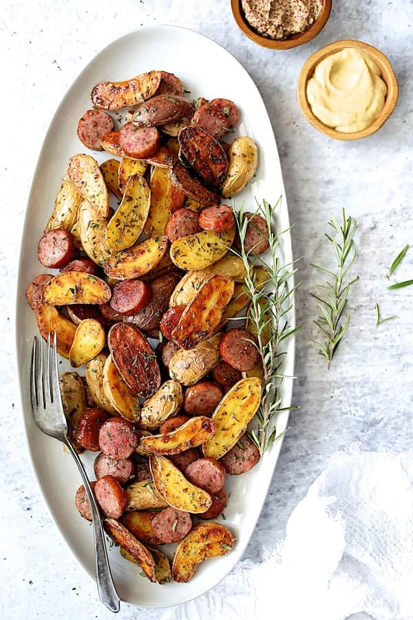 Photo of Duck Fat and Rosemary Roasted Fingerling with Sausage on white oval platter garnished with fresh rosemary.