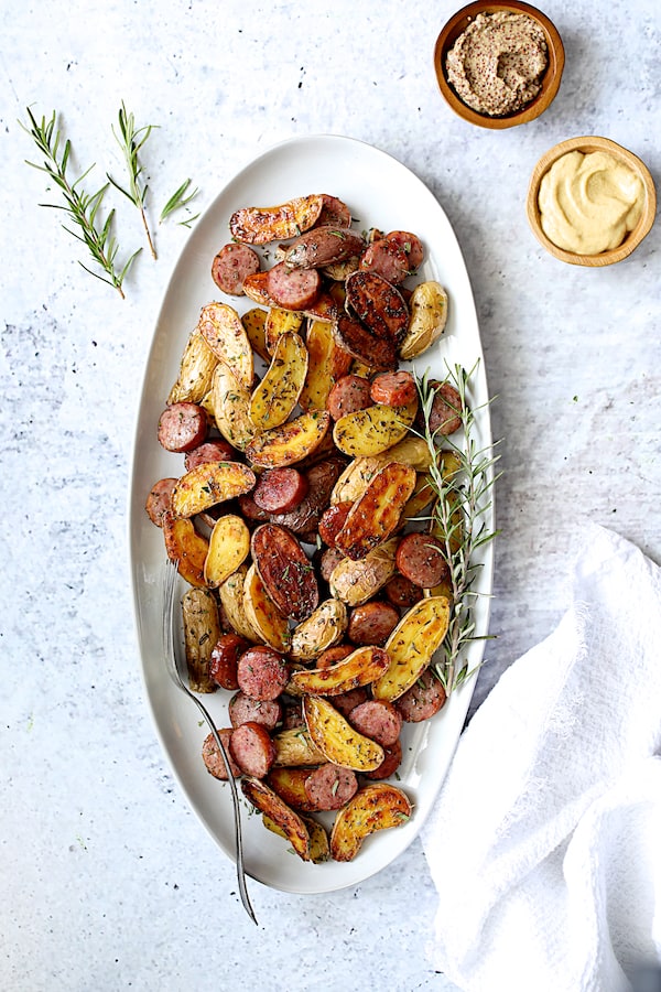 Photo of Duck Fat and Rosemary Roasted Fingerling Potatoes with Sausage on white oval platter with rosemary sprigs and bowls of mustards.