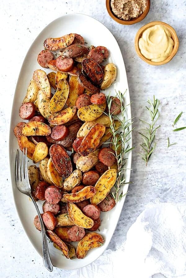 Duck Fat Roasted Fingerlings with Sausage on oval white platter garnished with fresh rosemary.