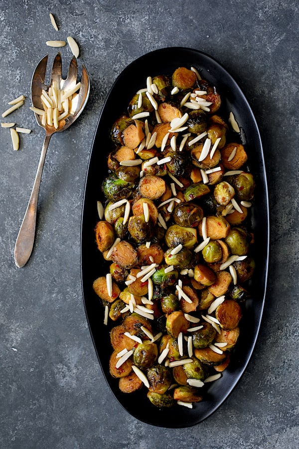 Photo of Pan-Roasted Brussels Sprouts with Thai Sweet Chili Orange Glaze on black serving platter with almonds scattered over.