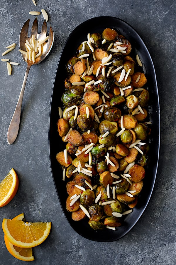 Photo of Pan-Roasted Brussels Sprouts with Thai Sweet Chili Orange Glaze on black platter with orange wedges.