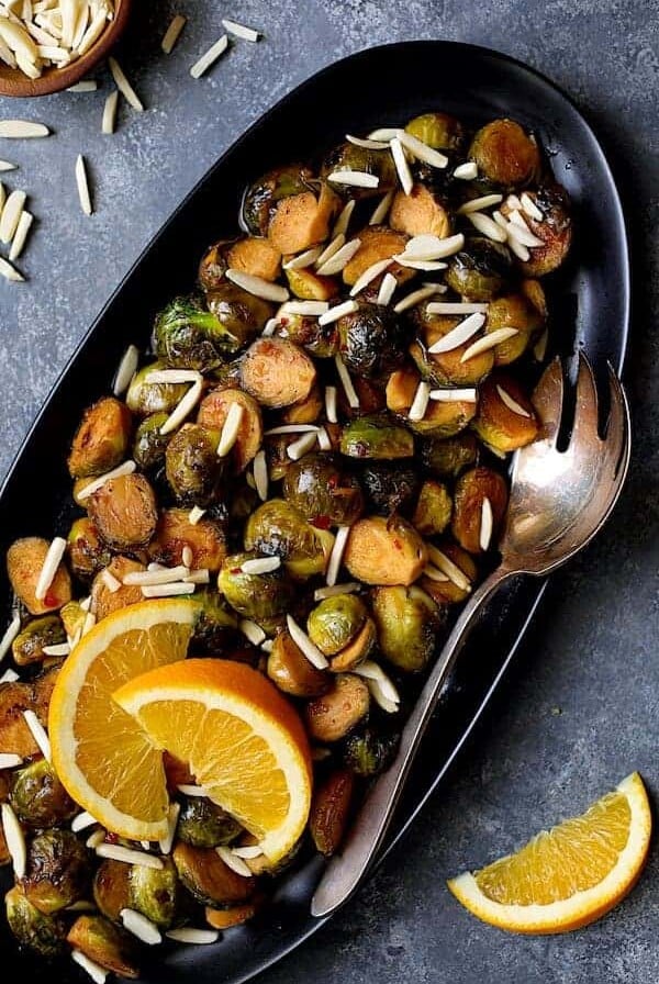 Pan-Roasted Brussels Sprouts with Thai Sweet Chili Orange Glaze on black oval platter with serving fork garnished with almonds and orange wedges.
