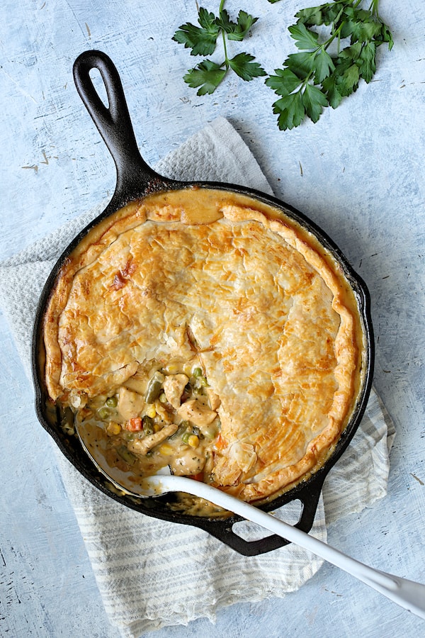 Photo of Cheesy Skillet Chicken Pot Pie on light blue background in cast iron skillet with some served to expose the filling.