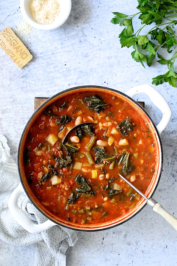 Photo of finished Italian Vegetable Stew in white Dutch oven on light blue background with cheese and parsley.