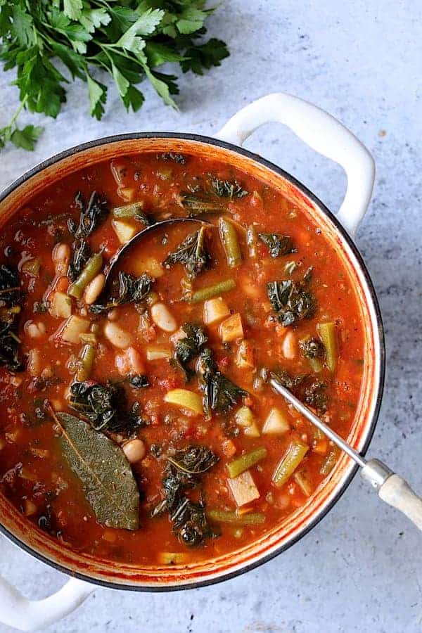 Photo of Italian Vegetable Stew in white Dutch oven with soup ladle on pale blue background.