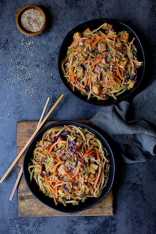 Thai Sesame Noodles with Tofu and Vegetables - Overhead shot of noodles in black bowls on dark blue background with chopsticks and gray napkin