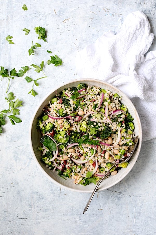 Photo of Lemony Broccoli, Barley and Bean Salad in serving bowl with parsley scattered about with white towel on pale blue background.