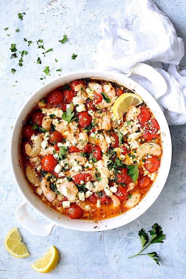 Photo of Spicy Baked Shrimp with Tomatoes and Feta Cheese on pale blue background with white towel, lemon wedges and parsley.