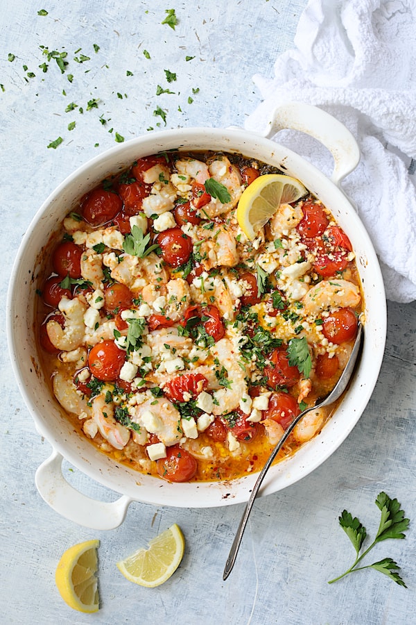 Photo of Spicy Baked Shrimp with Tomatoes and Feta Cheese on light blue background garnished with lemon and parsley.