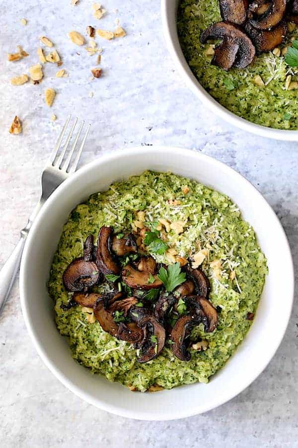 Photo of one serving of Oven Risotto with Kale Pesto and Roasted Mushrooms in white bowl on pale blue background.