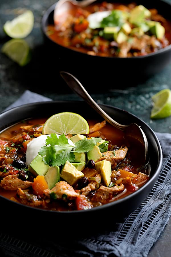 Slow Cooker Southwestern Turkey, Butternut Squash and Black Bean Stew - Straight-on shot of stew in black bowls on dark blue napkin garnished with lime, avocado and sour cream
