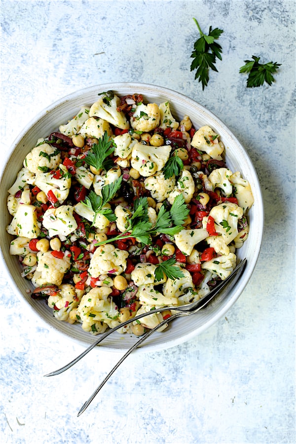 Photo of Sicilian Cauliflower Salad in white serving bowl with serving utensils garnished with parsley sprigs.