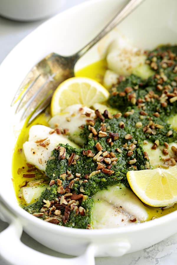 Close-up photo of Baked Tilapia with Spinach Pecan Pesto in white baking dish garnished with chopped pecans and lemon wedges.