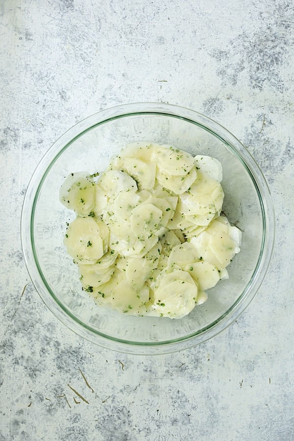 Photo of potato slices in glass bowl tossed with butter, oil and fresh herbs.