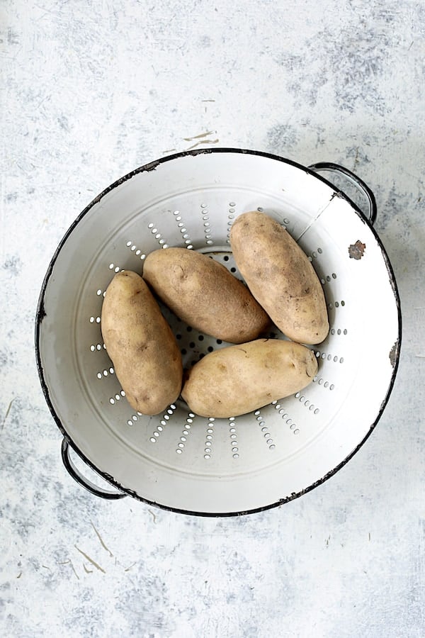 Photo of four unpeeled Russet potatoes in antique white colander.