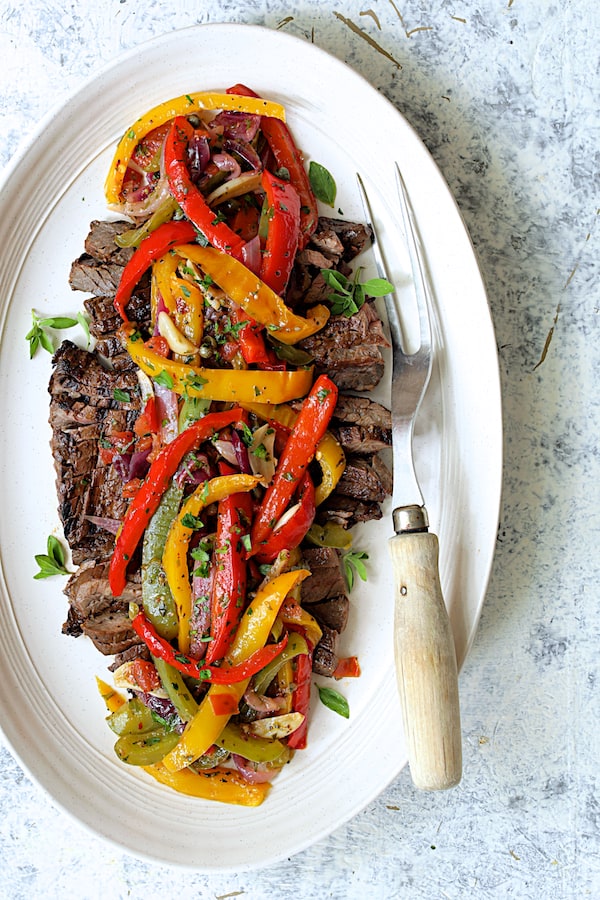 Photo of Grilled Marinated Skirt Steak with Peperonata on white oval platter on pale blue background garnished with fresh oregano.