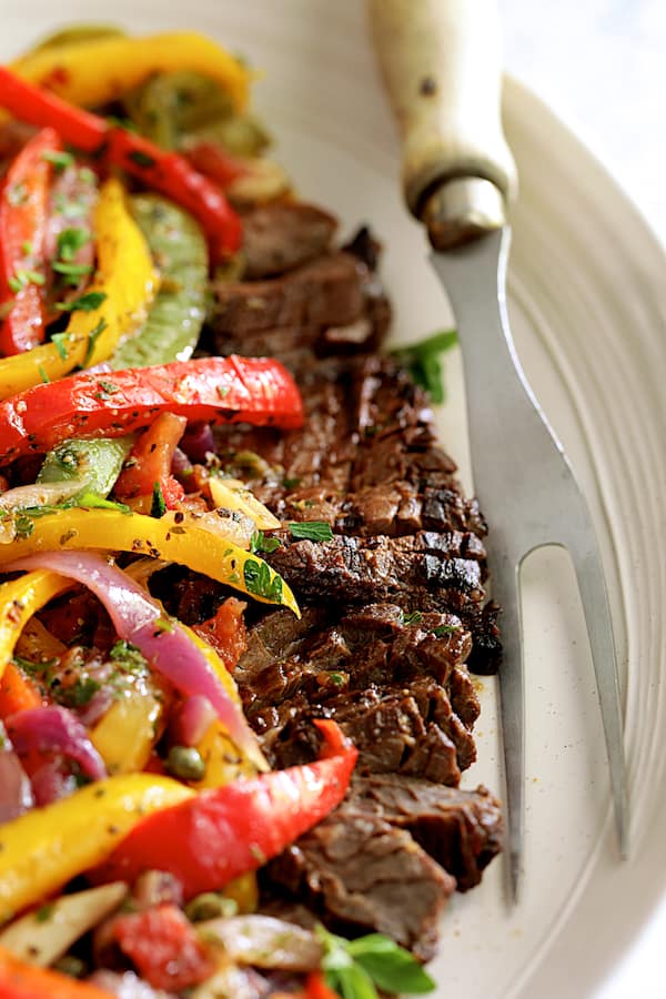 Close-up photo of Grilled Marinated Skirt Steak with Peperonata on white oval platter with meat fork.