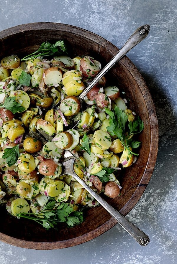 French-Style Potato Salad in wooden bowl with serving utensils garnished with fresh parsley.