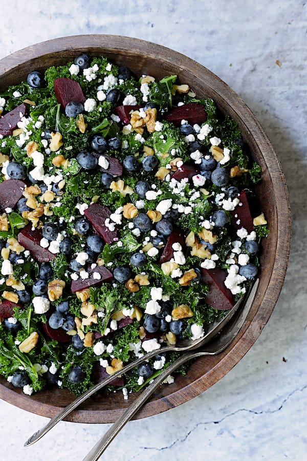 Photo of Massaged Kale, Beet and Blueberry Salad with Honey - Balsamic Dressing in wood bowl with serving utensils.