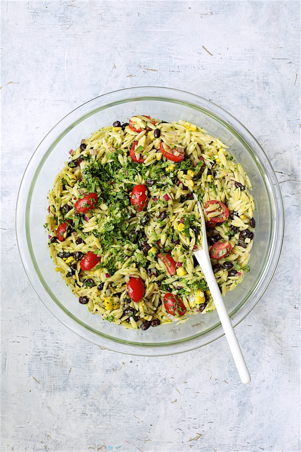 Photo of orzo salad being stirred together in glass bowl with white spoon.