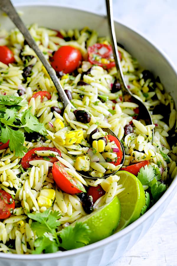 Close-up photo of Southwestern Orzo Salad with Cilantro Lime Dressing in white serving bowl with utensils.