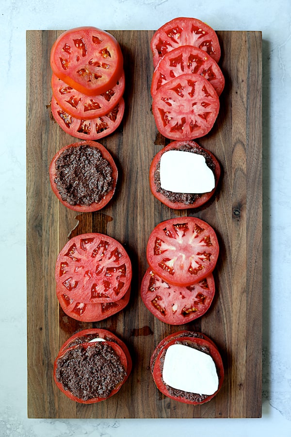 Photo of tomato slices on wood cutting board being layered with tapenade and fresh mozzarella.