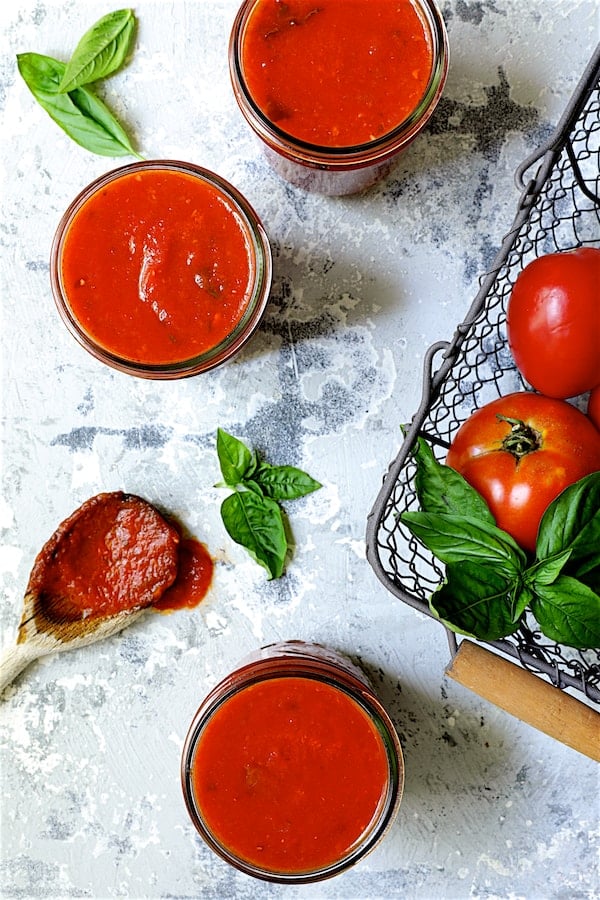 Photo of three jars of Basic Fresh Tomato Sauce (Parma Style) in pint jars with tomatoes in a basket and sauce on wooden spoon.