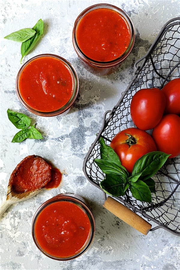 Photo of three jars of Basic Fresh Tomato Sauce (Parma Style) in glass pint jars with tomatoes in wire basket and some sauce on a spoon.