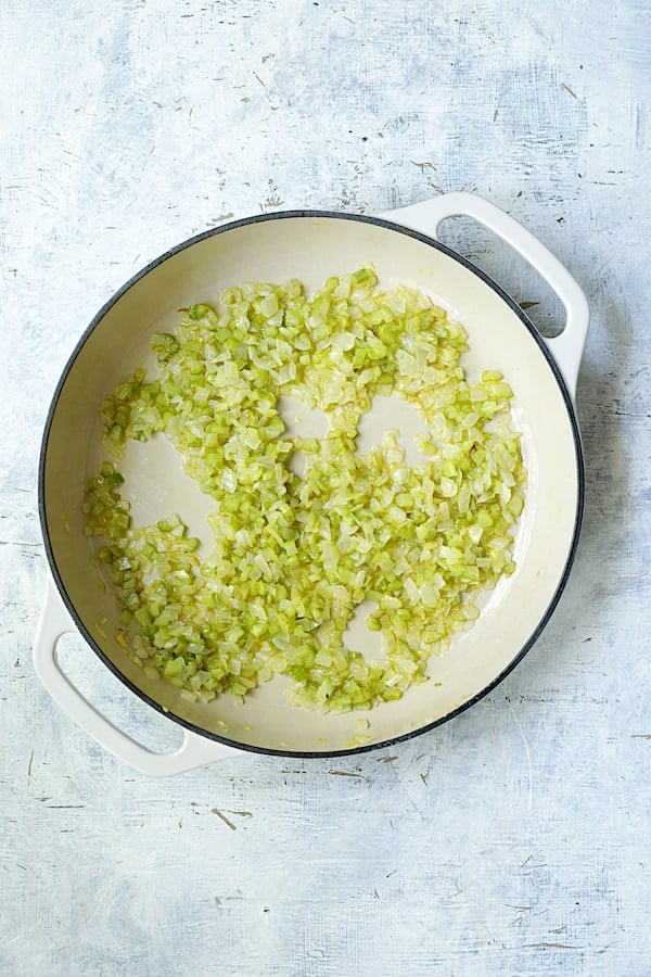 Photo of onion and celery being cooked in white cast iron skillet.