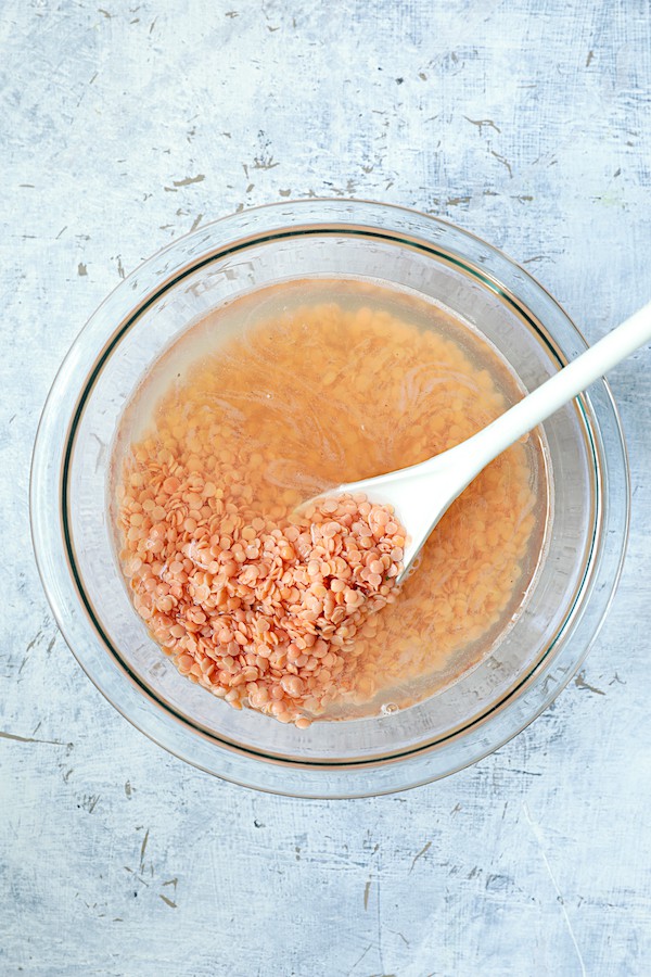 Photo of red lentils being soaked in glass bowl and being stirred with white spoon.