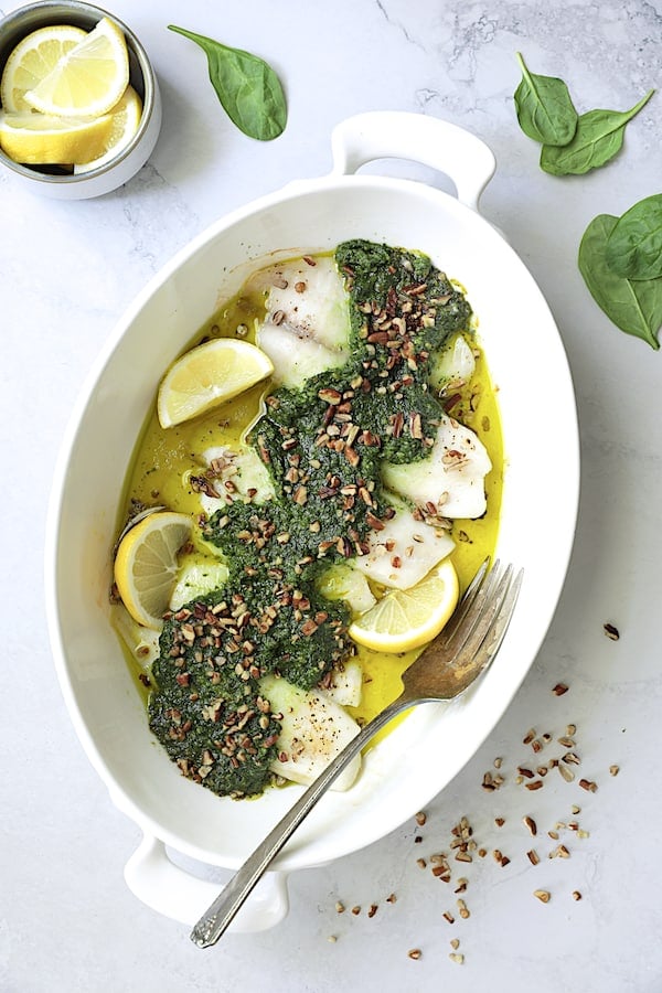 Photo of Baked Tilapia with Spinach Pecan Pesto on light gray background garnished with pecans and lemon wedges.