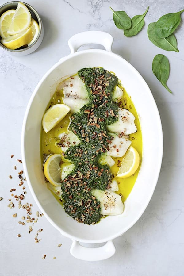 Photo of Baked Tilapia with Spinach Pecan Pesto in white oval baking dish garnished with lemon wedges and chopped pecans.