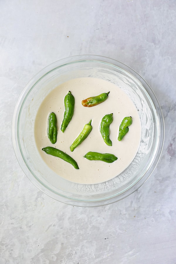 Photo of shishito peppers being dipped into batter in glass bowl.