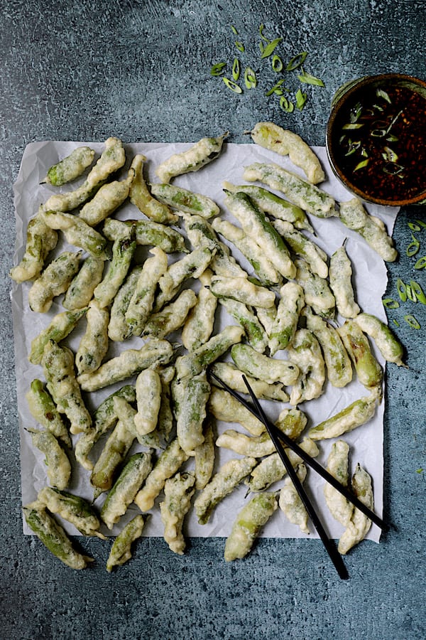Photo of of tempura shishito peppers on white parchment paper with black chopsticks.