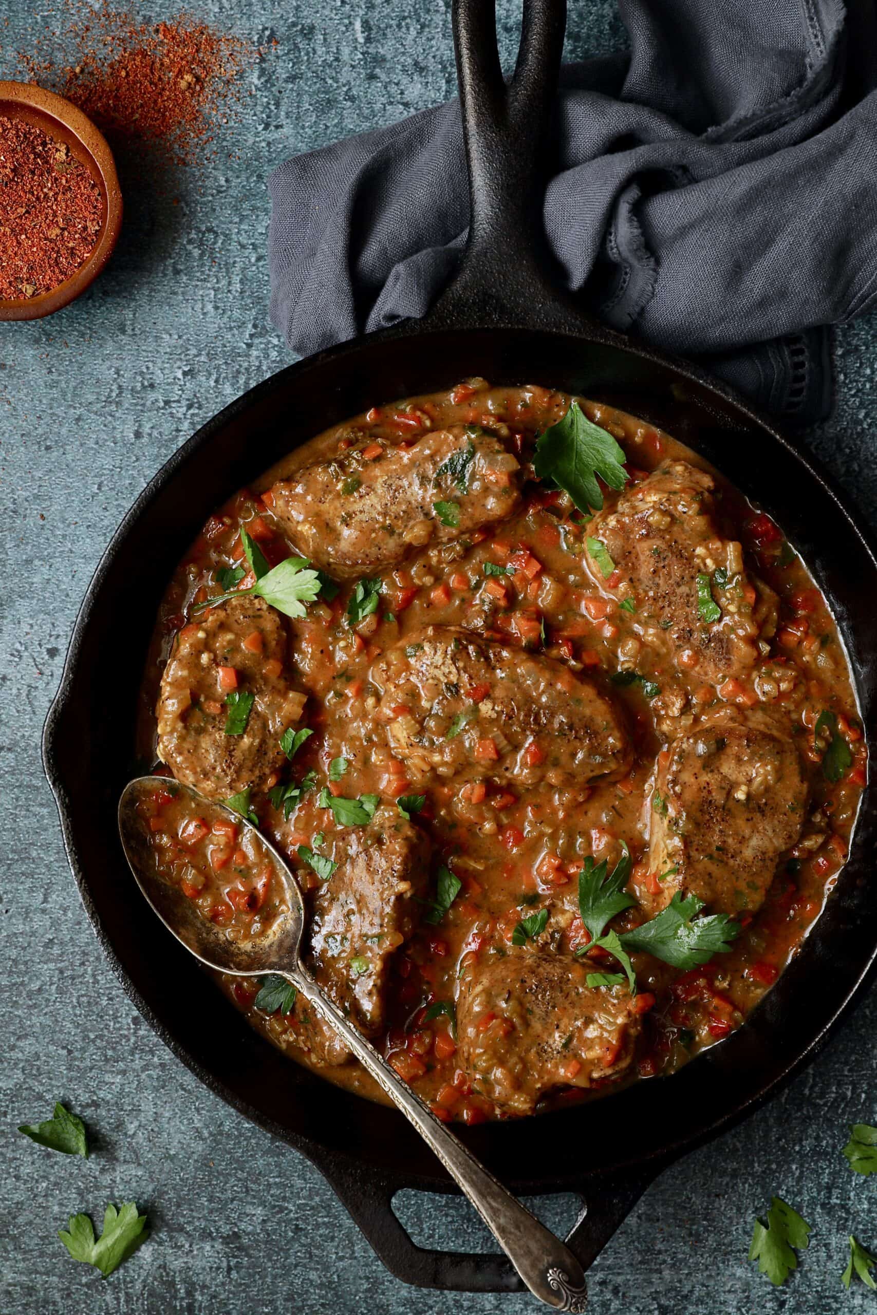 Photo of Cajun-Smothered Pork Medallions in cast iron skillet on blue background.