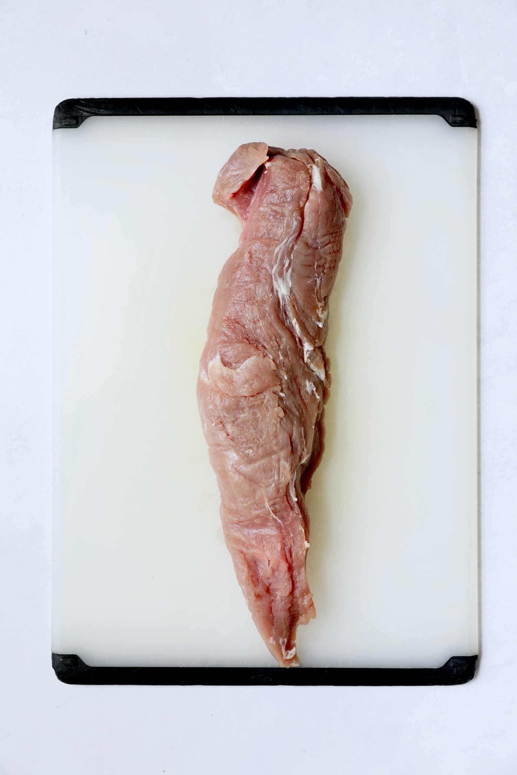 Photo of whole uncooked pork tenderloin on white cutting board.