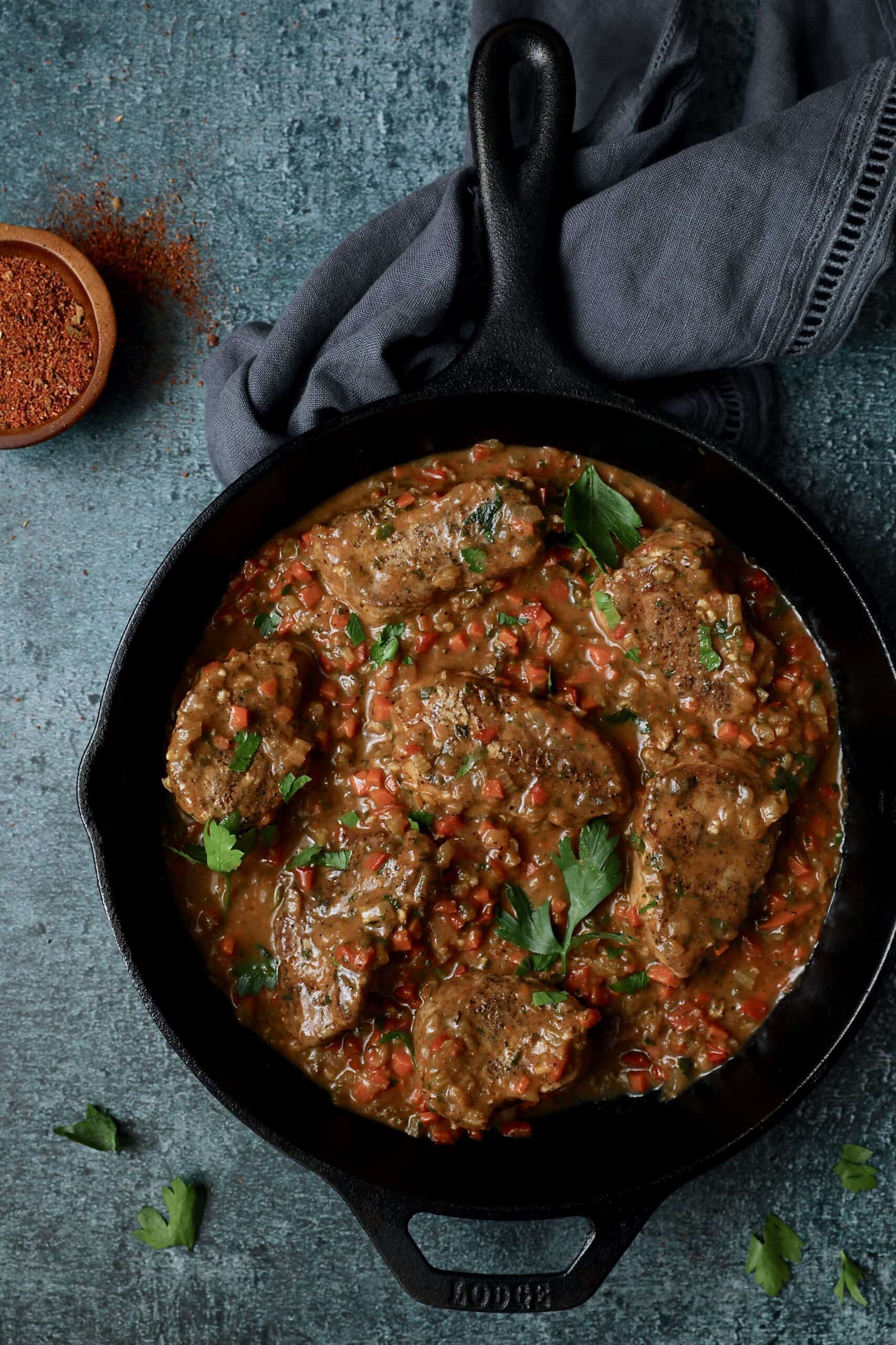 Photo of Cajun-Smothered Pork Medallions in cast iron skillet on blue background garnished with parsley.