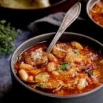 Chorizo White Bean and Vegetable Soup - Straight-on close-up shot of soup in gray bowl with soup spoon and bread in the background