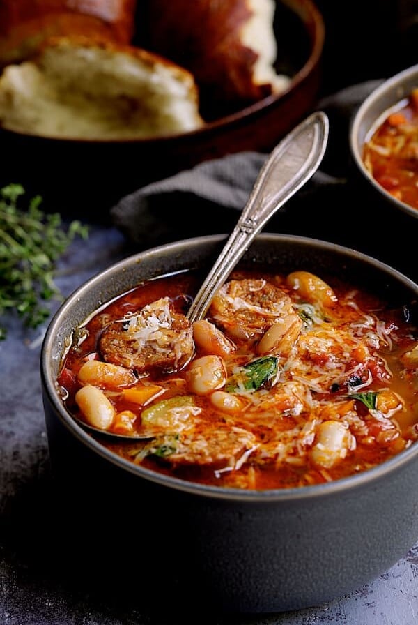 Chorizo White Bean and Vegetable Soup - Straight-on close-up shot of soup in gray bowl with soup spoon and bread in the background