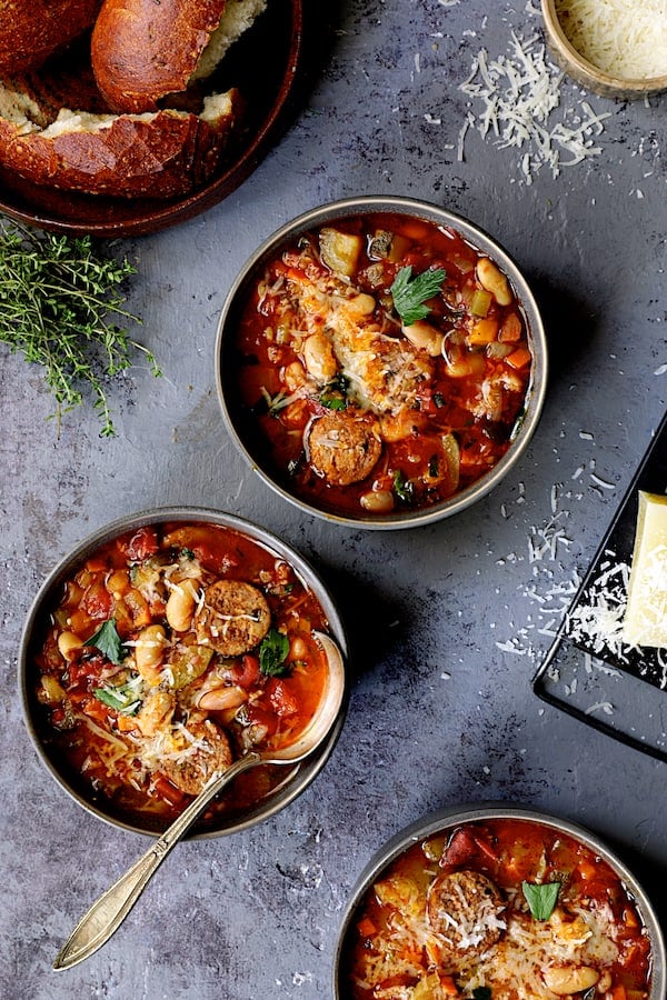Photo of Chorizo White Bean and Vegetable Soup in gray bowls with bread and cheese sprinkled about.