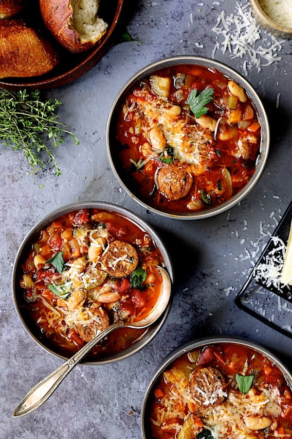 Photo of three bowls of Chorizo White Bean and Vegetable Soup in gray bowls on gray background with cheese and bread.