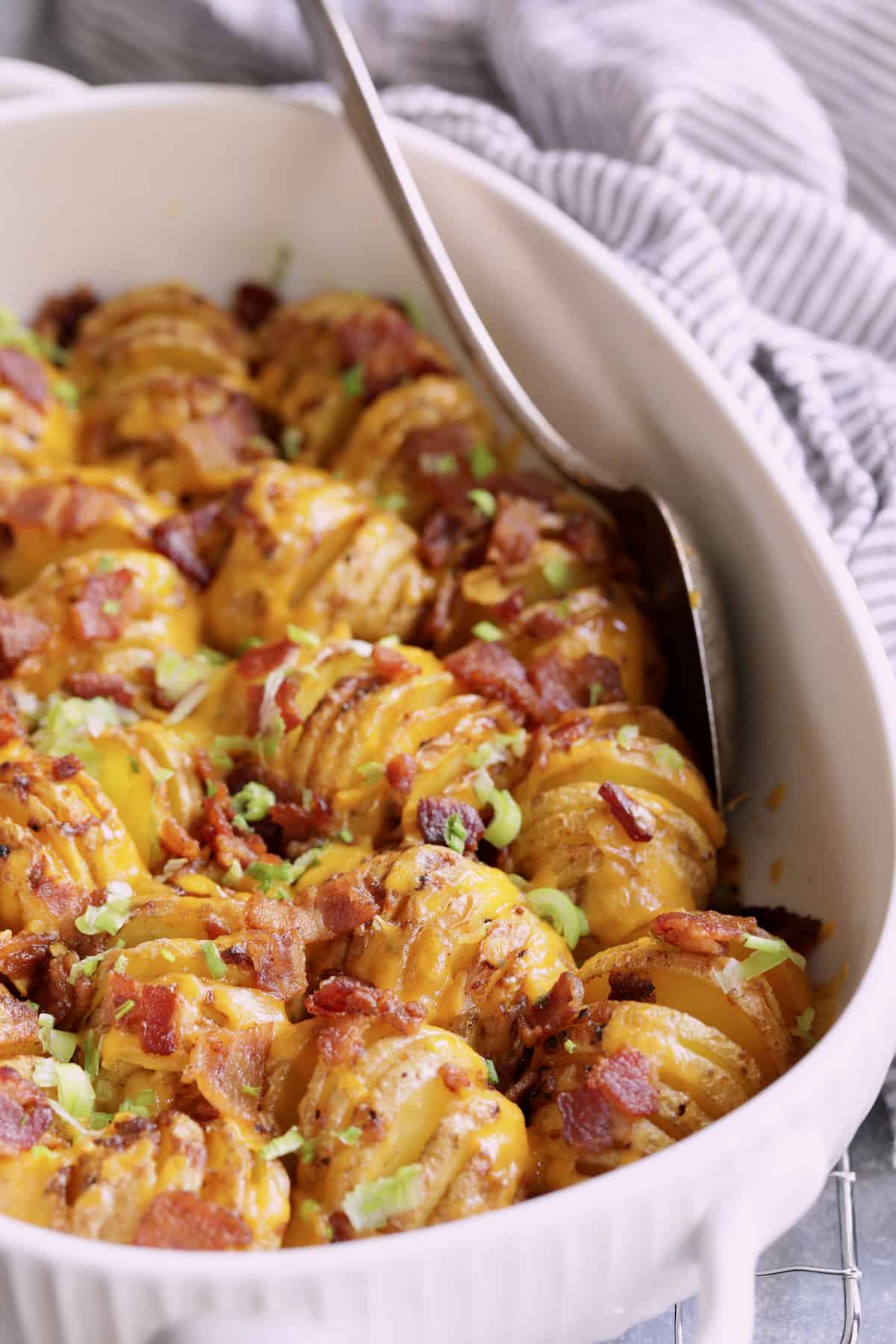Close-up photo of Baby Hasselback Potato Bake with Cheddar and Bacon in white baking dish with serving spoon.