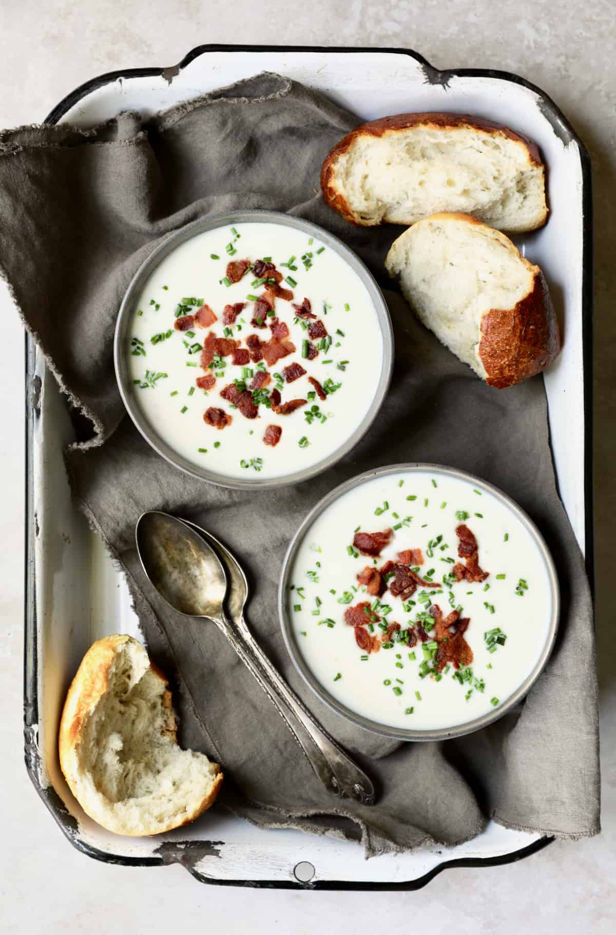 Photo of two bowls of Potato Leek Soup with White Cheddar on gray napkin in white tray with bread.