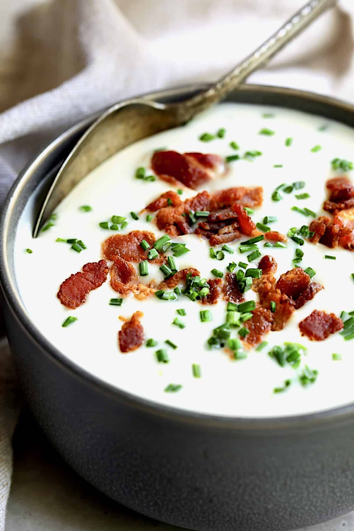 Close-up photo of Potato Leek Soup with White Cheddar in gray bowl with spoon garnished with bacon and chives.
