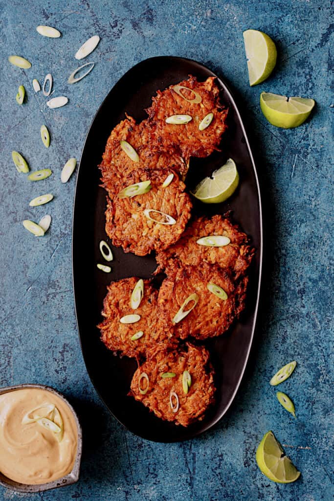 Photo of Korean Sweet Potato Latkes with Gochujang Mayonnaise on black platter on bright blue background garnished with lime wedges and scallion.