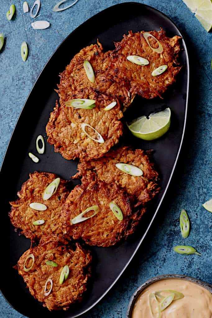 Photo of Korean Sweet Potato Latkes with Gochujang Mayonnaise on black platter garnished with lime wedges and scallion.