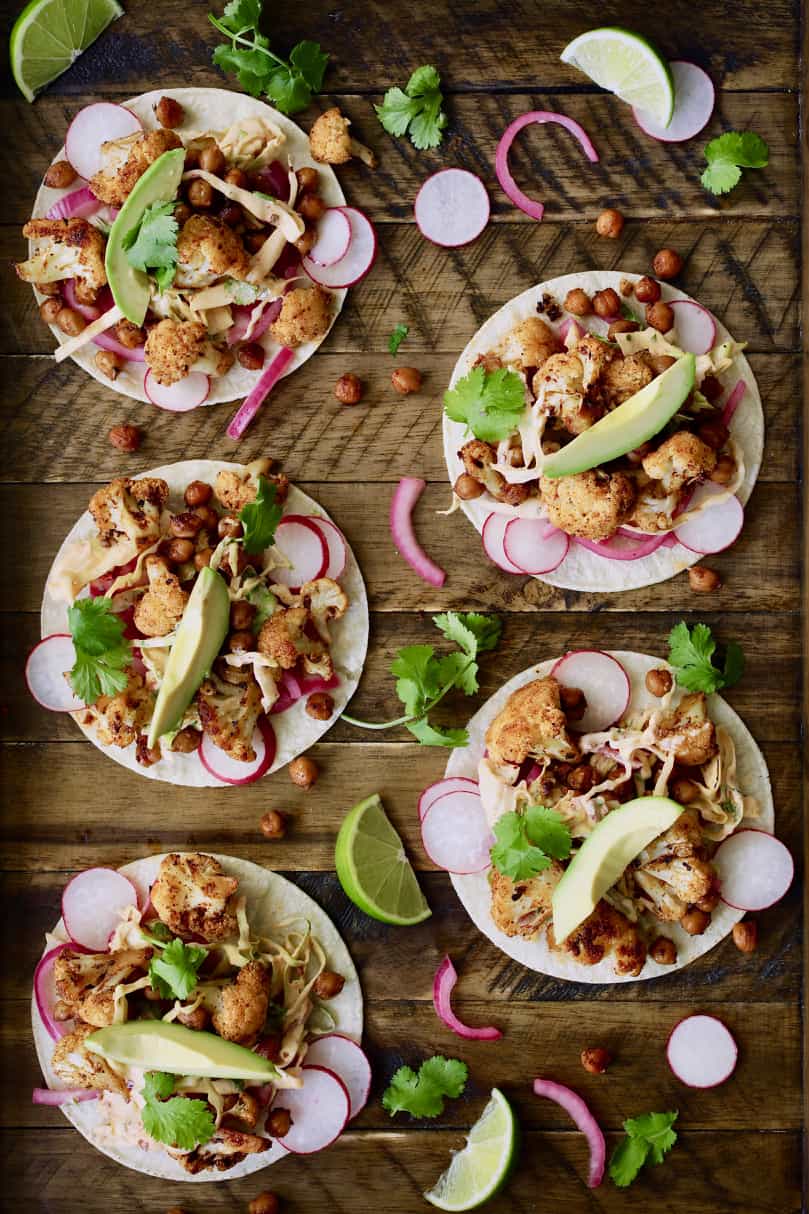 Photo of Cauliflower and Chickpea Tacos with Chipotle Lime Slaw on wood board garnished with radishes, avocado, onions and lime wedges.