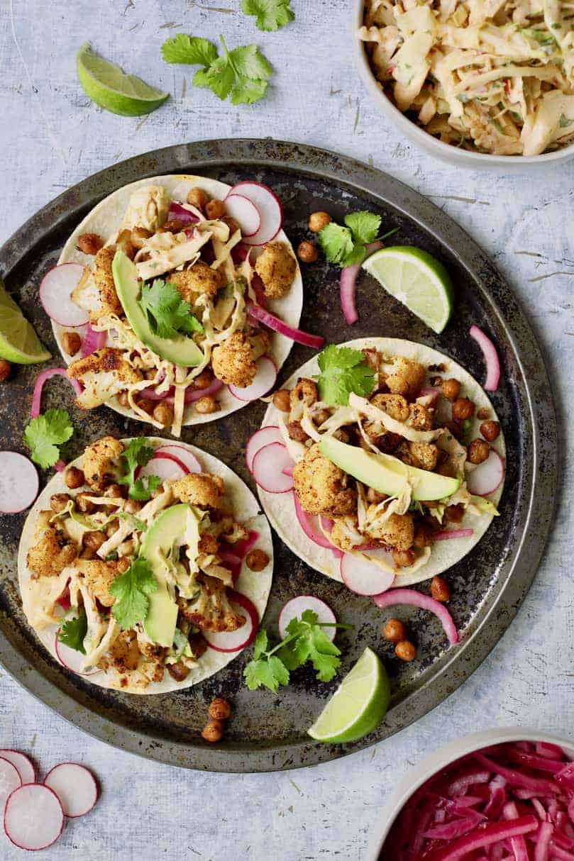 Photo of three Cauliflower and Chickpea Tacos with Chipotle Lime Slaw with garnishes on light blue background.